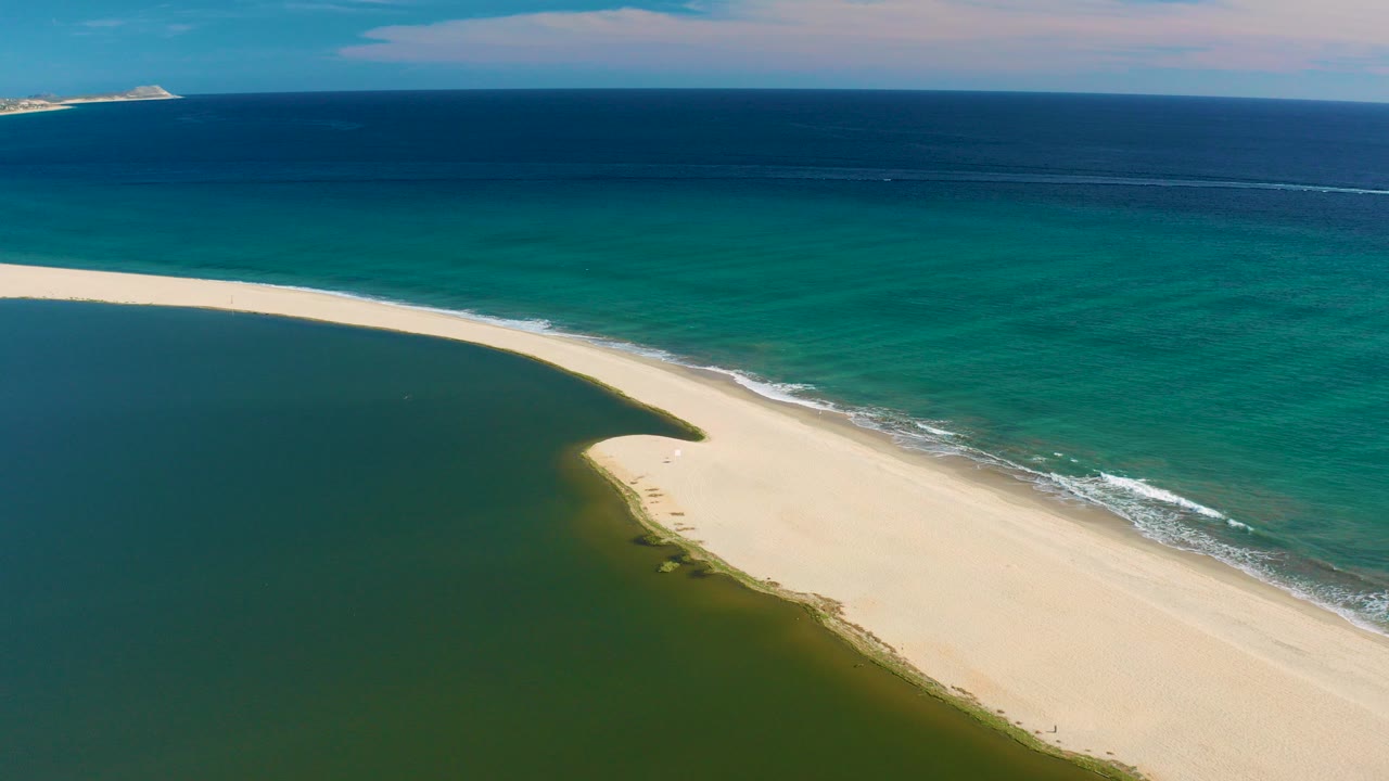 Aerial frame from San Jose Del Cabo showing a pale sand barrier separating dark lagoon water from turquoise ocean, with white surf on the beach and no visible people.