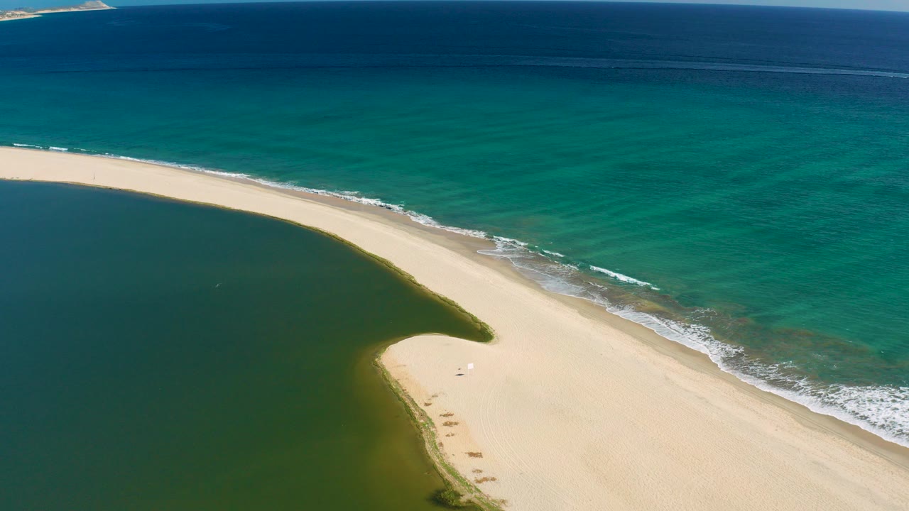 At 0:00, an aerial view in San Jose del Cabo shows a pale curved sandbar separating dark green lagoon water from turquoise ocean, with no visible people.