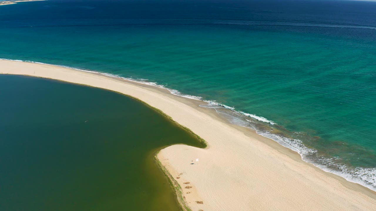 At 0:02, the empty San Jose del Cabo beach strip cuts diagonally across the frame, with darker lagoon water below and clear turquoise ocean above, with no visible people.
