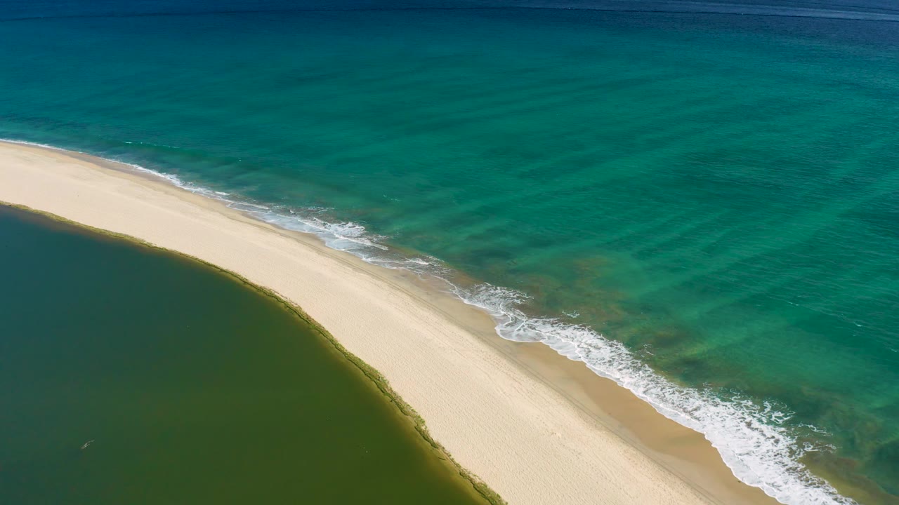 A diagonal sandbar in San Jose Del Cabo separates the stiller green lagoon from the rippled turquoise ocean, with surf foam shifting along the right shoreline and no visible people.
