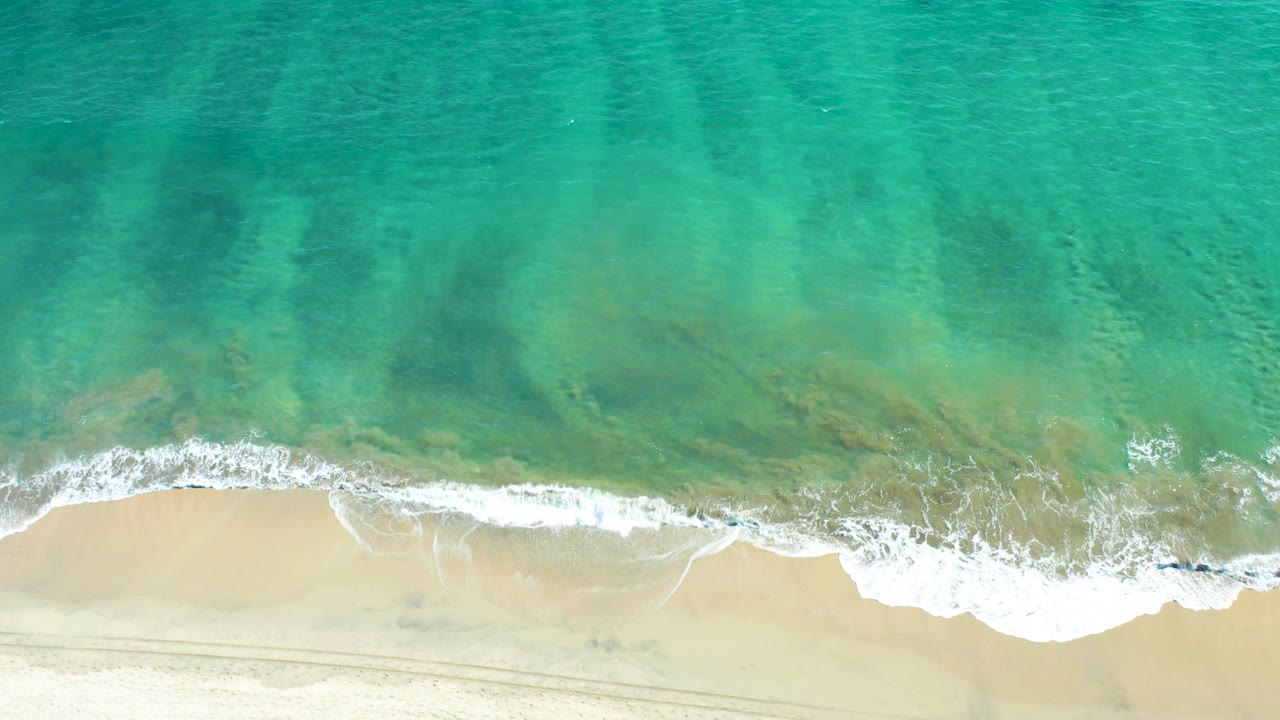 Overhead view of turquoise water washing onto pale sand in San Jose Del Cabo, Mexico, with a thin line of white foam and no visible people.