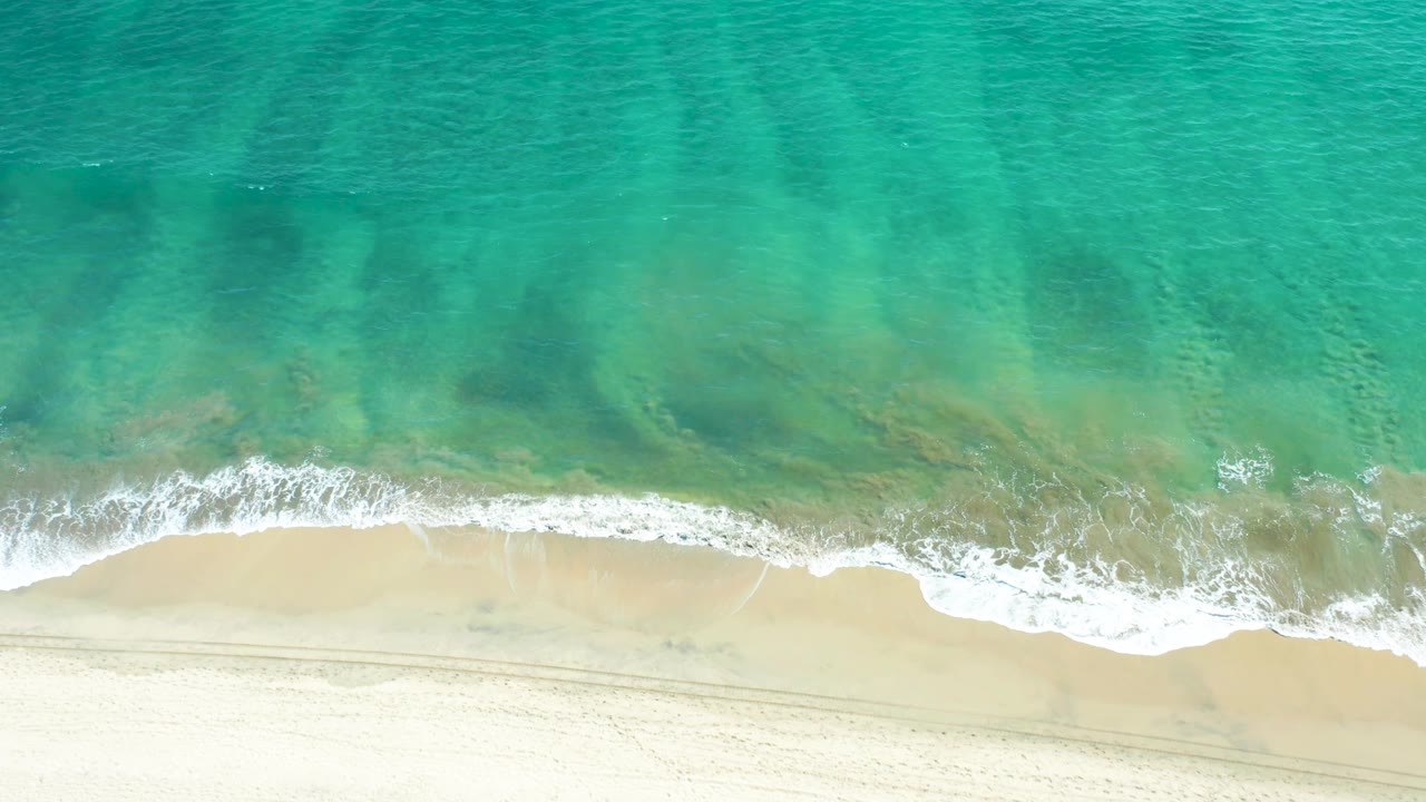 The overhead view continues across San Jose Del Cabo beach, where turquoise water meets a wider pale sand band marked by faint parallel tracks and no visible people.