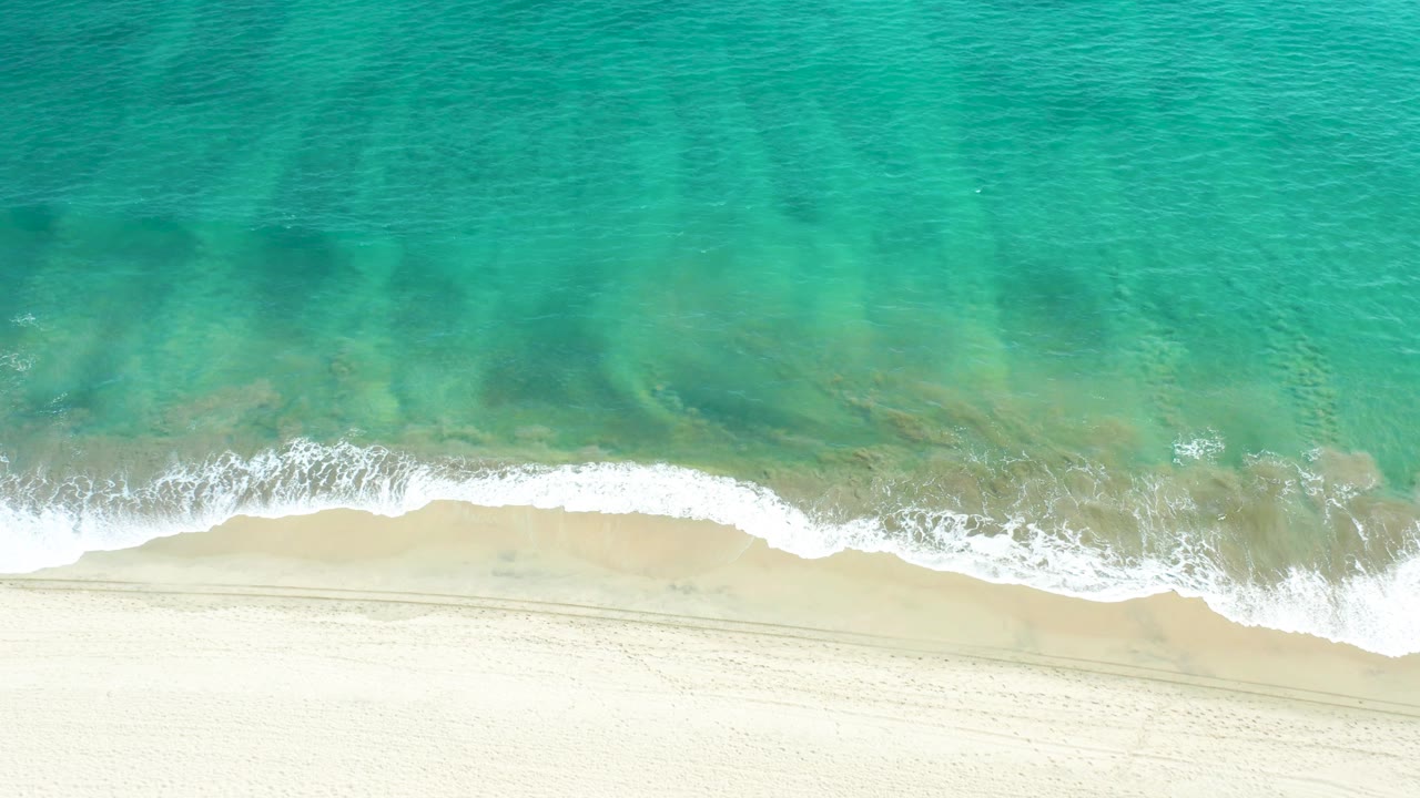 Mid-clip overhead view of San Jose Del Cabo surf, with clear turquoise water covering most of the frame, a narrow white foam line, pale sand below, and no visible people.