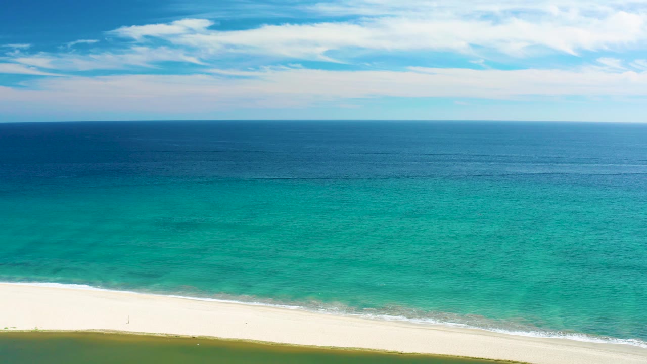 Mid-clip San Jose Del Cabo shoreline view with white sand crossing the lower frame, turquoise water beyond it, light cloud cover overhead, and no visible people.