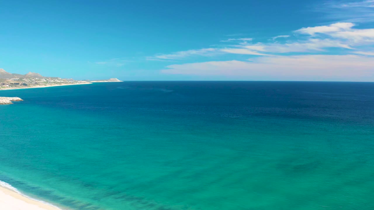 Opening frame of San Jose Del Cabo turquoise shallows, deep blue ocean, pale sand at the bottom edge, distant left coastline, and no visible people.