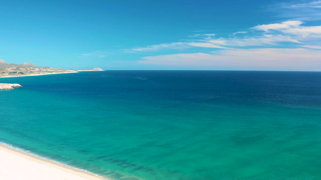San Jose del Cabo beach aerial with a wider white sand edge at bottom left, layered turquoise water, darker offshore ocean, pale clouds, and no visible people.