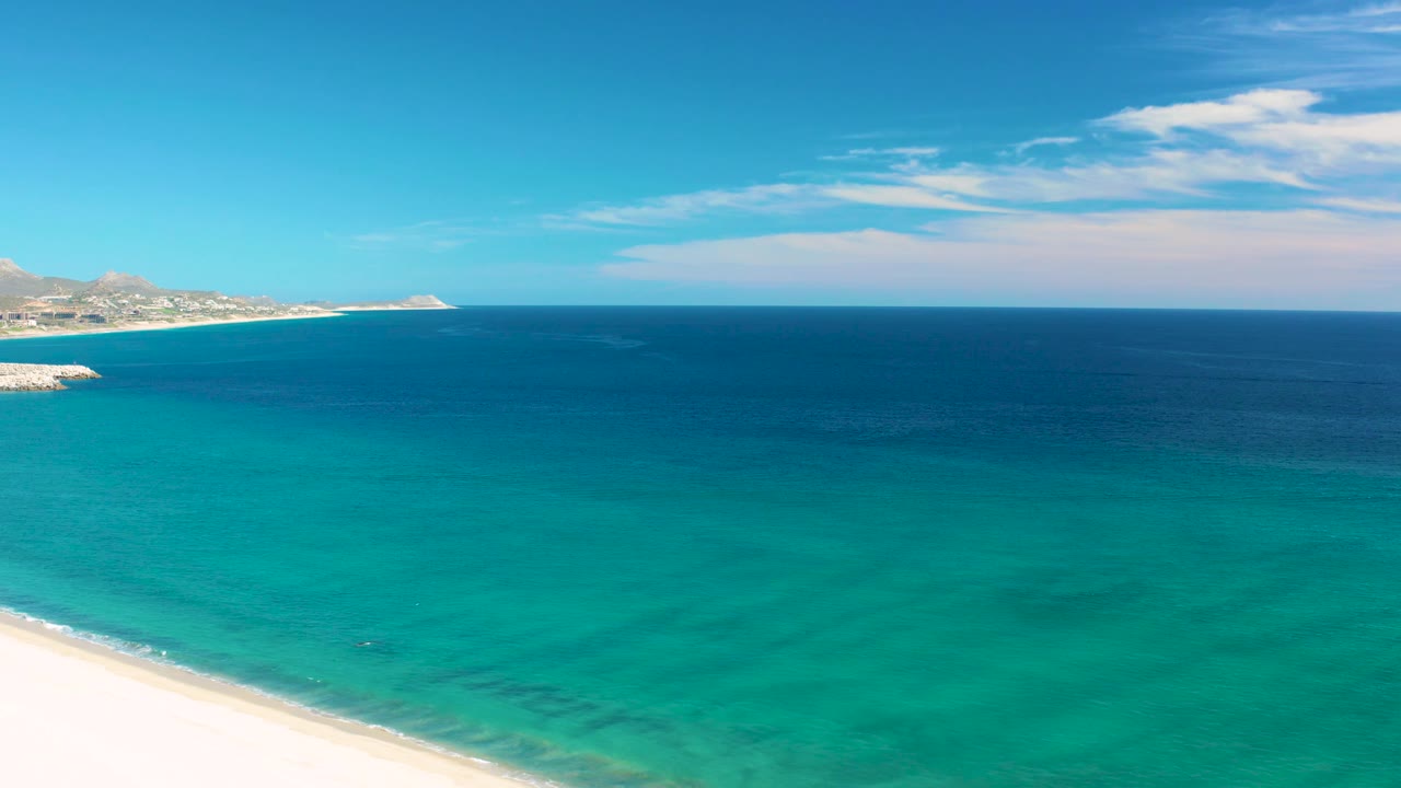 San Jose del Cabo shoreline with bright sand along the bottom, clear turquoise shallows filling the frame, dark blue sea beyond, distant land at left, and no visible people.