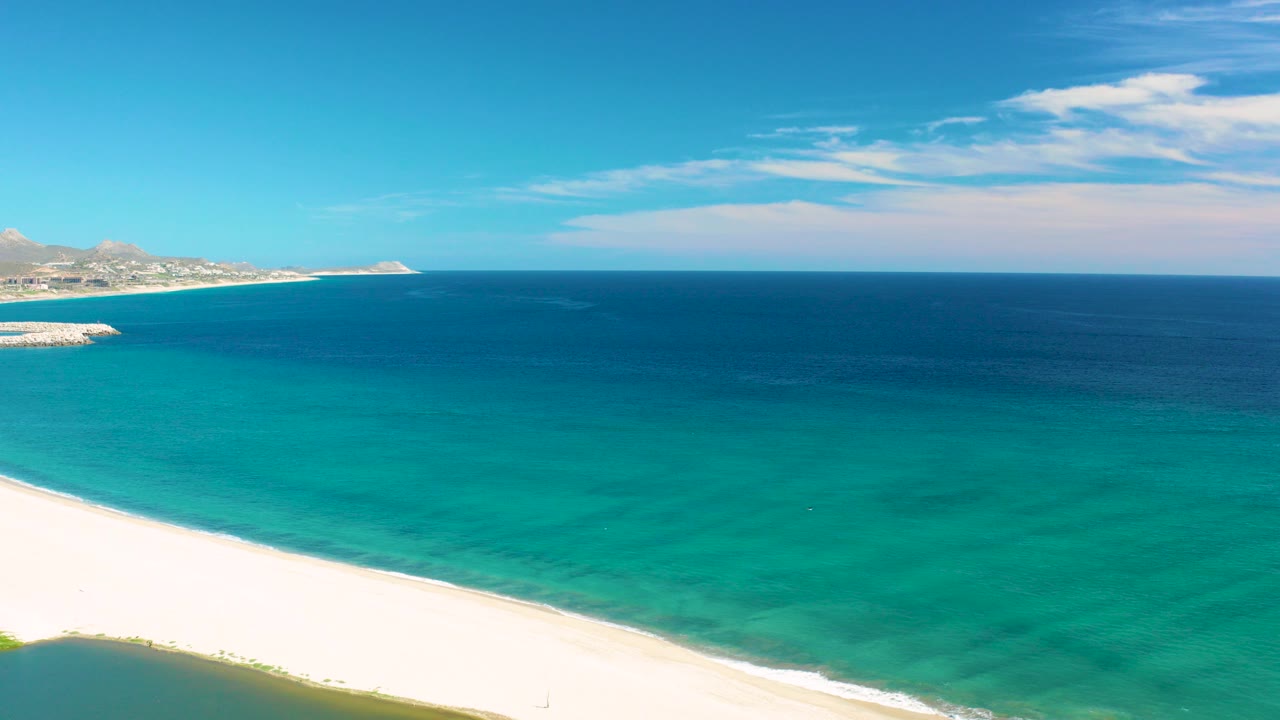 Aerial view in San Jose del Cabo, Mexico, with white sand along the bottom edge, turquoise shallows, deep blue ocean, distant coastline on the left horizon, and no visible people.