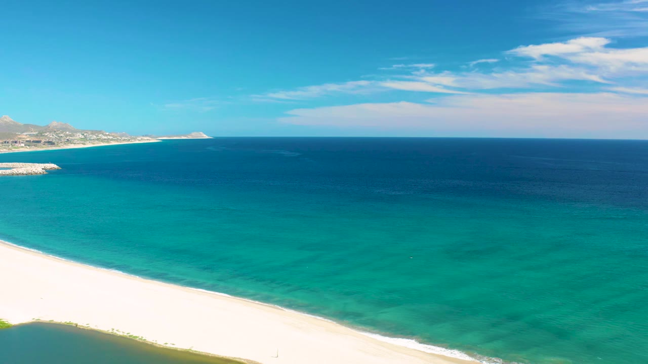 Aerial coastal frame in San Jose del Cabo, Mexico, showing a bright sand strip, transparent turquoise water, darker ocean beyond, a low horizon, and no visible people.