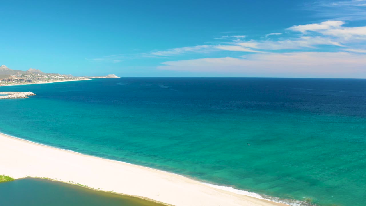 San Jose del Cabo beach seen from above, with the sand line bending across the lower frame, green-turquoise water near shore, open blue sea beyond, and no visible people.