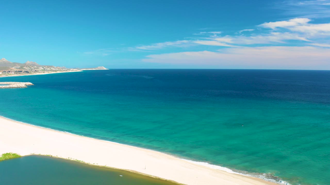 Elevated view over San Jose del Cabo coastline, with a white beach band in the foreground, turquoise water across the middle, distant built shoreline at left, and no visible people.