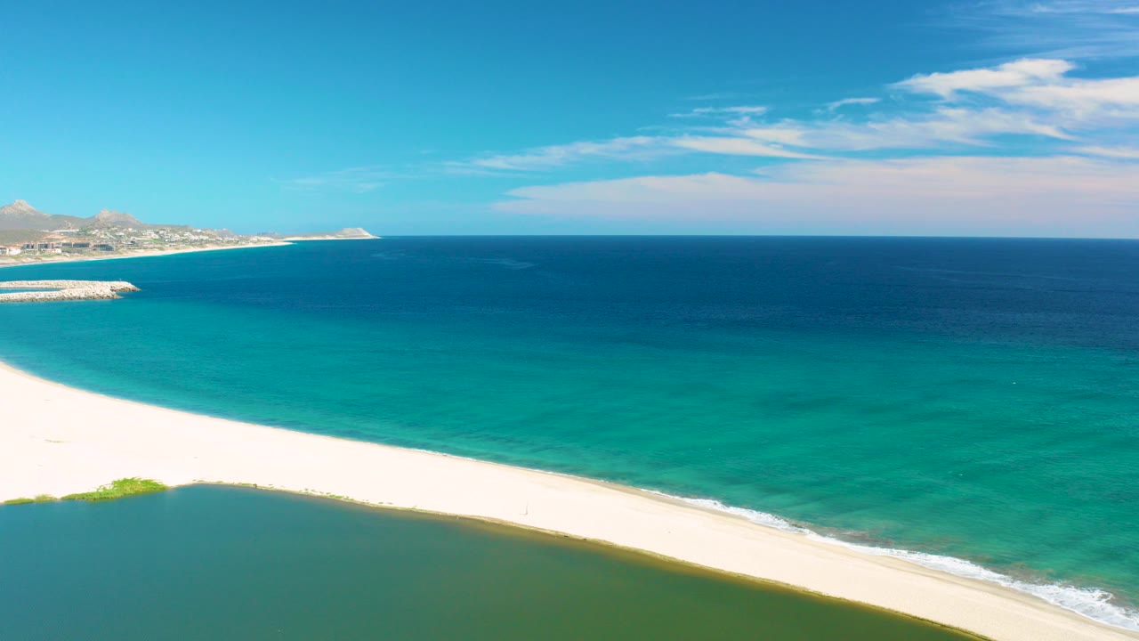 Aerial view in San Jose Del Cabo showing turquoise ocean beyond a white sand beach, a green-blue lagoon in the foreground, distant coastline at left, and no visible people.