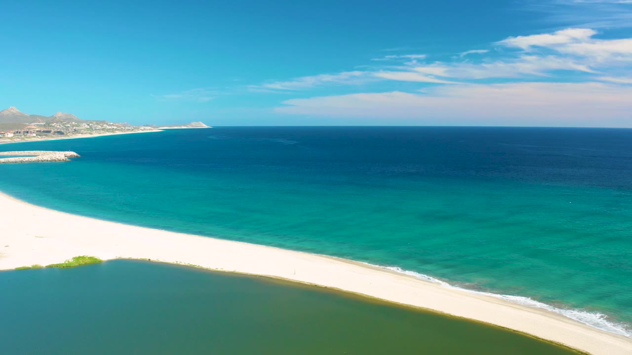 Wide aerial frame over San Jose Del Cabo with lagoon water filling the lower frame, white shoreline sweeping across the middle, deep blue ocean above, and no visible people.