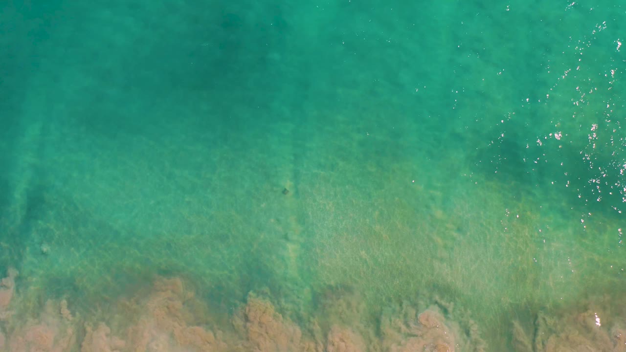 Top-down view of turquoise water over a sandy seabed in San Jose del Cabo, with darker underwater bands and no visible people.