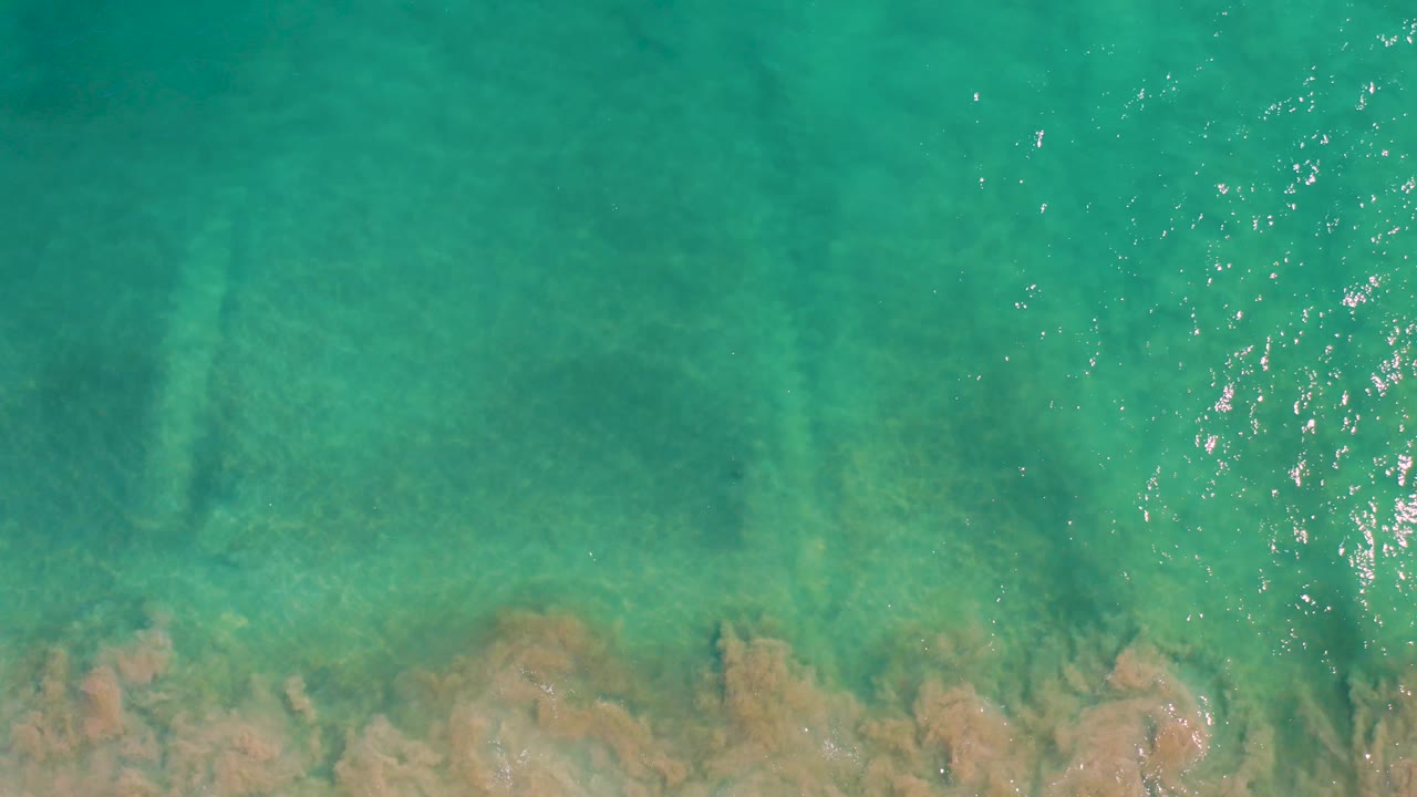 Top-down San Jose Del Cabo water frame with green-blue shallows, faint darker patches, tan rocks along the lower frame, and no visible people.