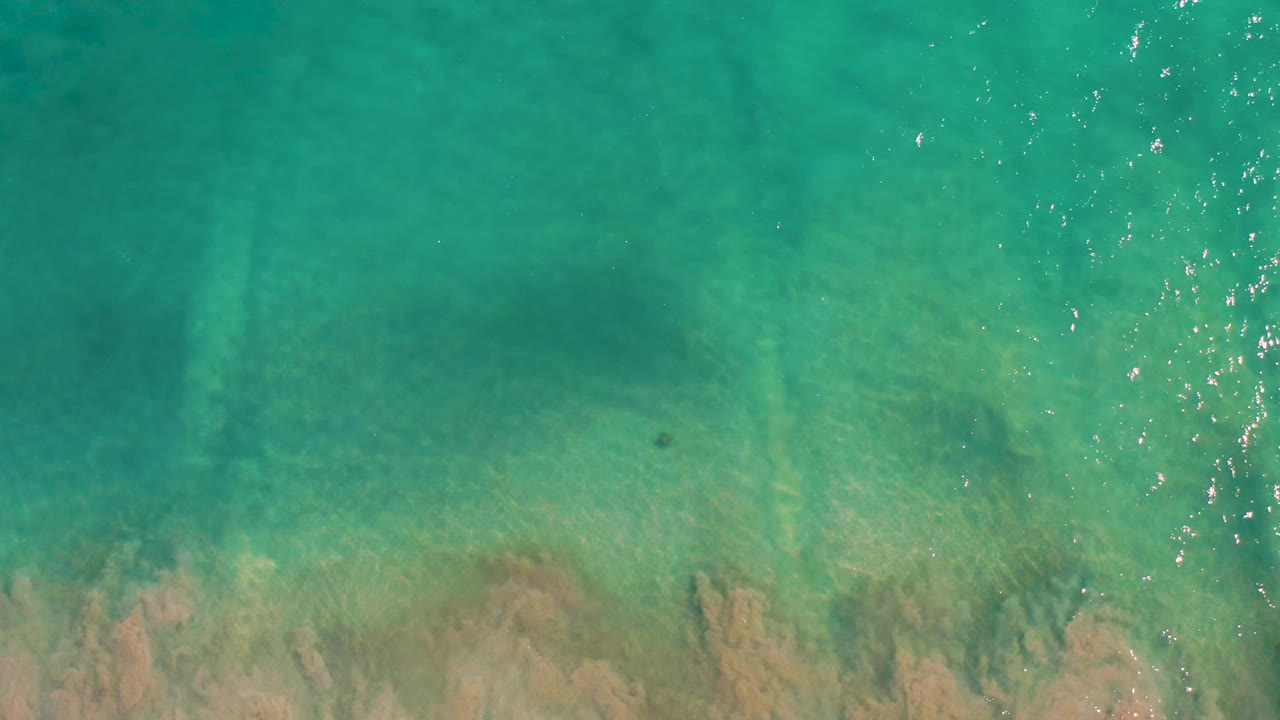 Clear turquoise water in San Jose Del Cabo shows a submerged tan rock formation near the lower frame and bright surface glints with no visible people.