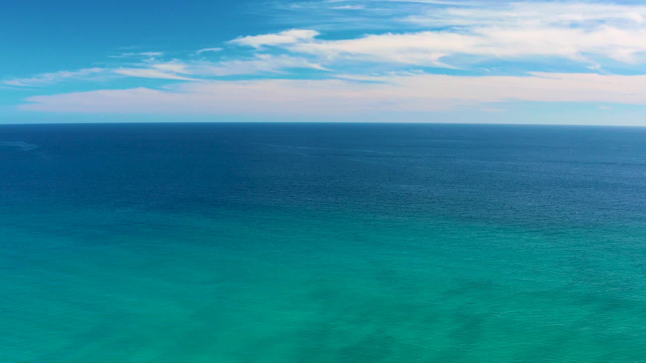 Aerial view over San Jose del Cabo ocean water, with turquoise water in the foreground, darker blue water near the horizon, scattered white clouds above, and no visible people.