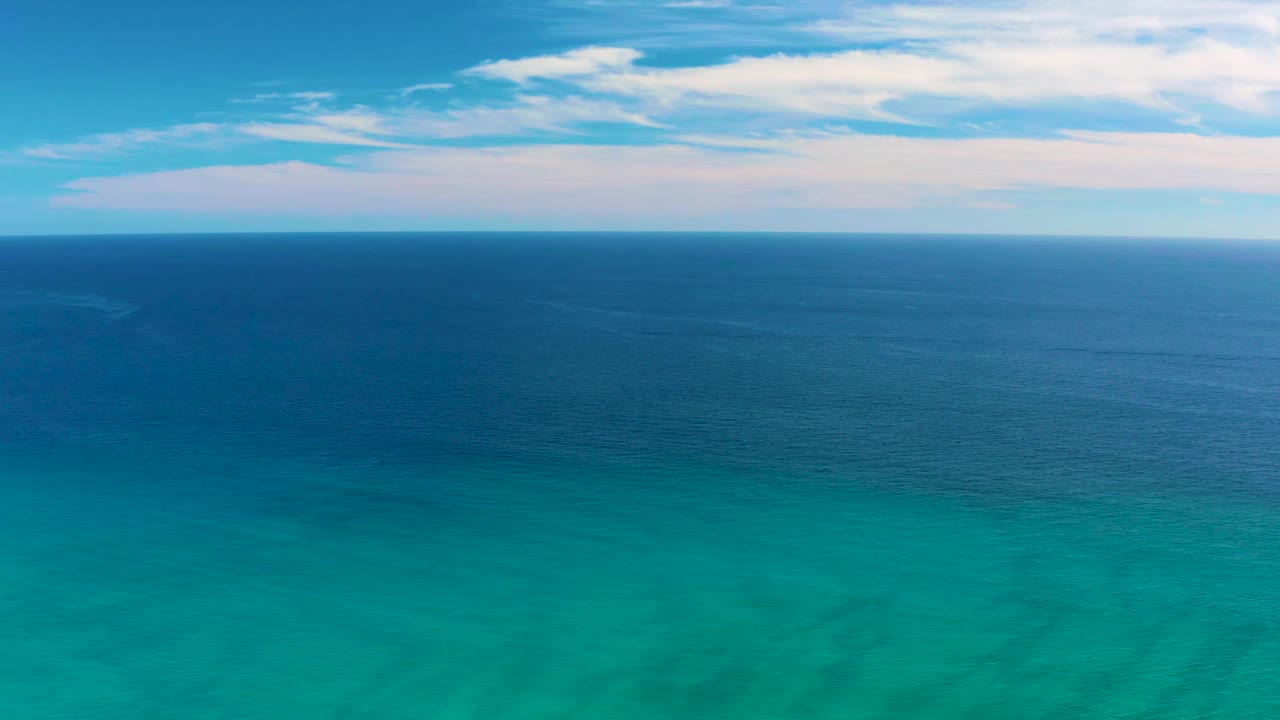 A steady aerial frame from San Jose del Cabo shows calm turquoise foreground water, a darker blue band of ocean, a level horizon, thin clouds, and no visible people.