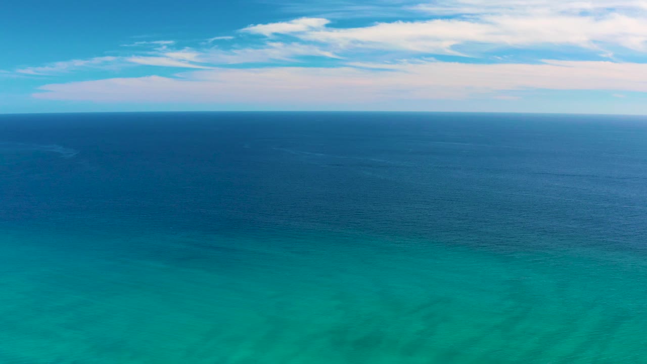 A wide San Jose del Cabo aerial frame holds on rippled turquoise water, a darker blue midground sea, pale sky, streaked white clouds, and no visible people.