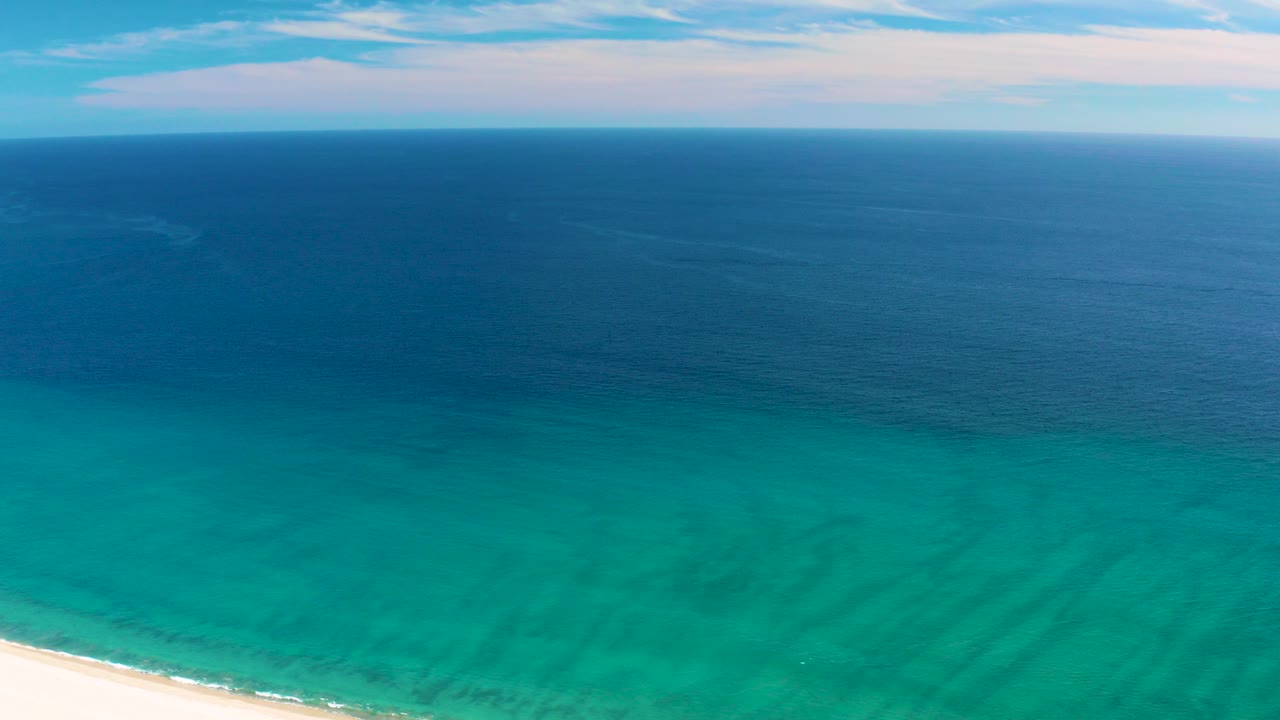 Opening frame of San Jose del Cabo coastline with white sand at the lower left, turquoise shallows, darker blue sea, pale clouds, and no visible people.