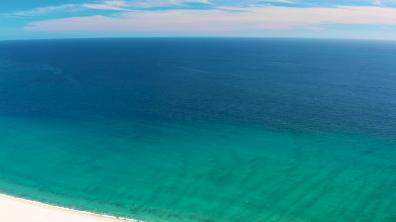 San Jose del Cabo beach curves across the lower frame with shallow turquoise water, darker ocean past the color break, thin clouds, and no visible people.