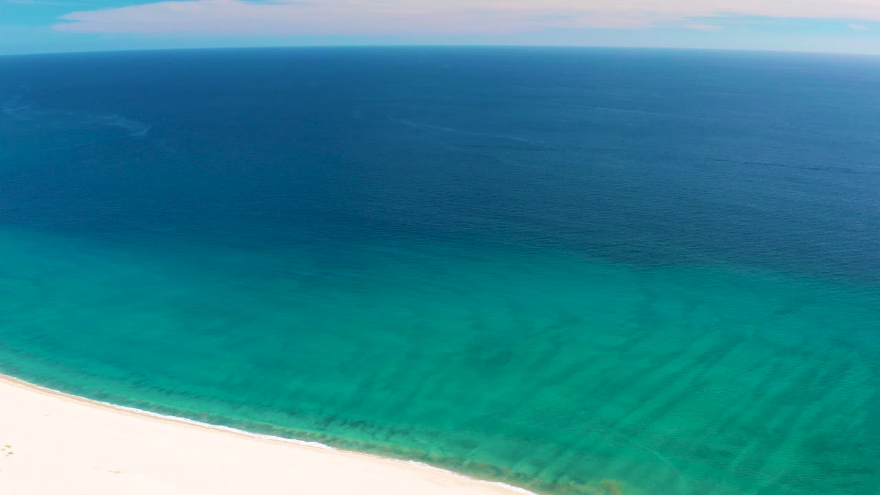Aerial view over San Jose del Cabo beach with white sand at the lower left, turquoise shallow water, deeper blue ocean beyond, and no visible people.