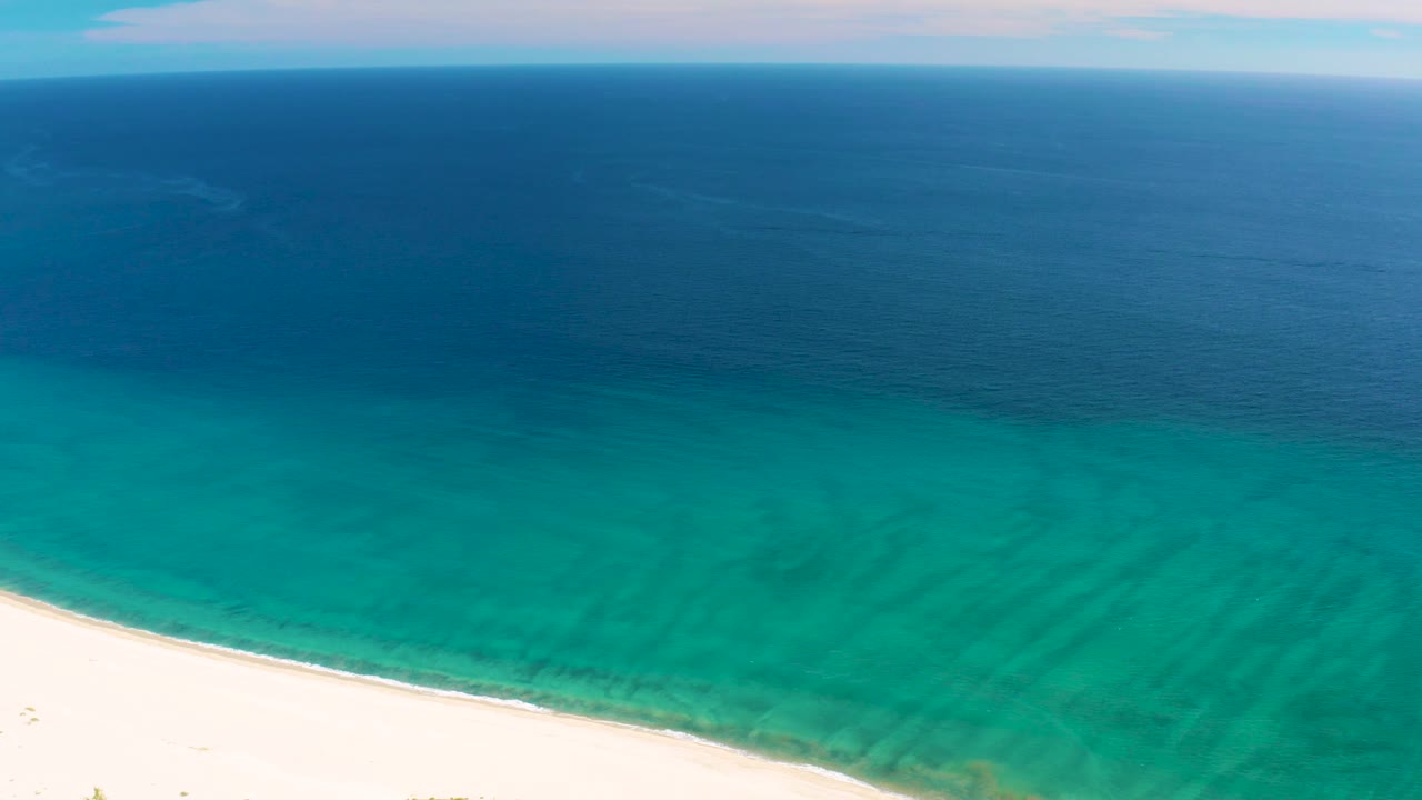 Aerial frame from San Jose del Cabo with a longer sweep of white shoreline at the bottom, textured turquoise water in the center, deep blue ocean above, and no visible people.
