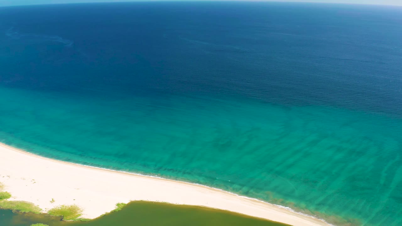 Aerial frame from San Jose del Cabo with the beach curving through the lower image, pale sand, green vegetation patches, layered turquoise water, and no visible people.