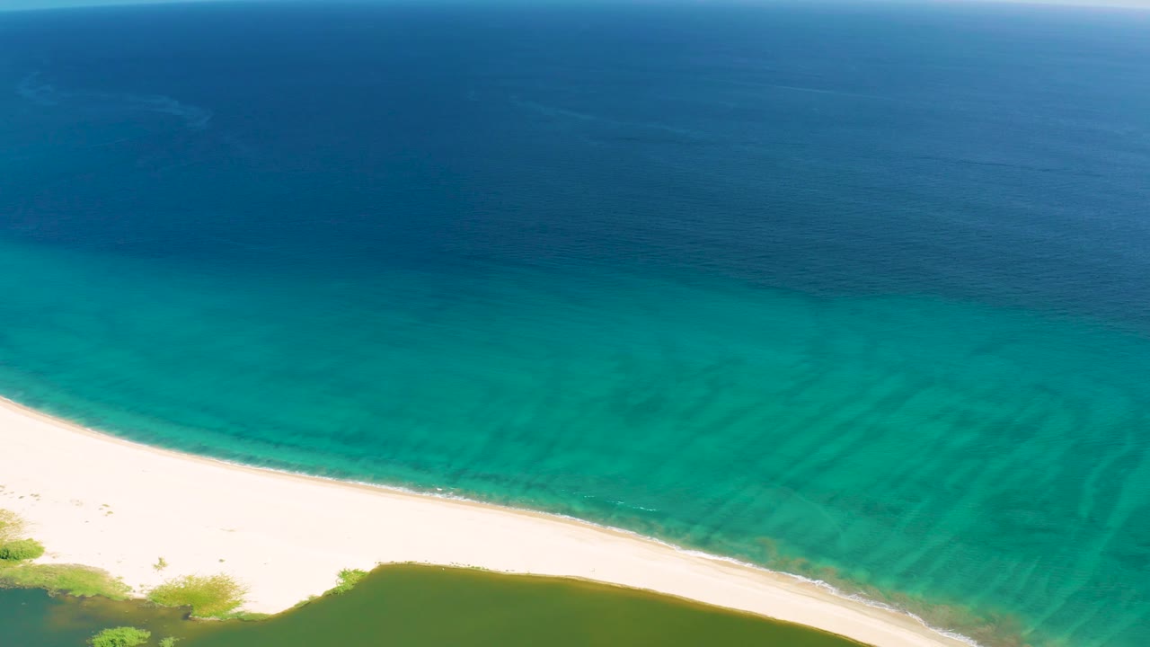Aerial frame from San Jose del Cabo showing a bright strip of white beach, green vegetation at the lower edge, turquoise nearshore water, deep blue ocean, and no visible people.