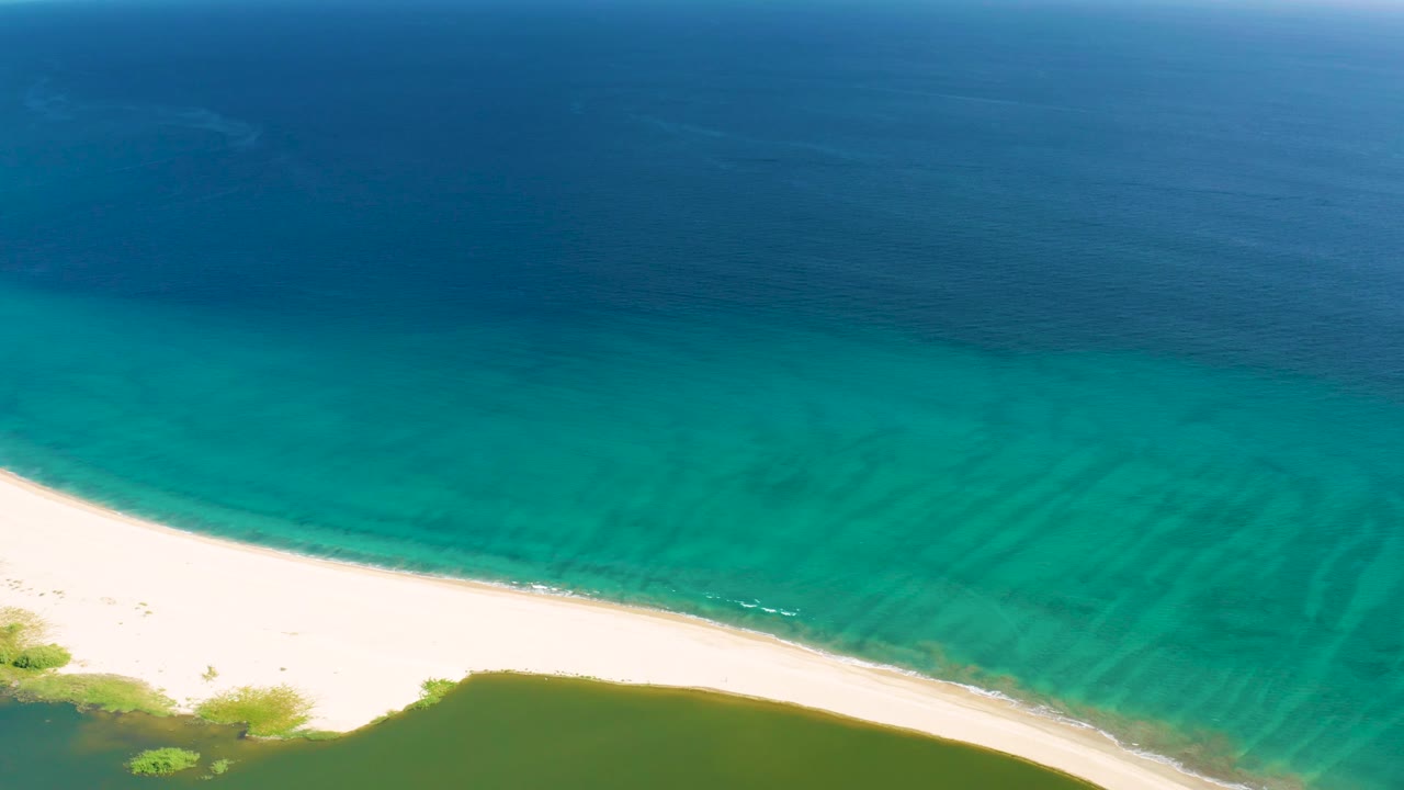 Aerial frame from San Jose del Cabo where pale sand runs along the bottom left, turquoise water spreads across the midframe, darker ocean sits above, and no visible people appear.