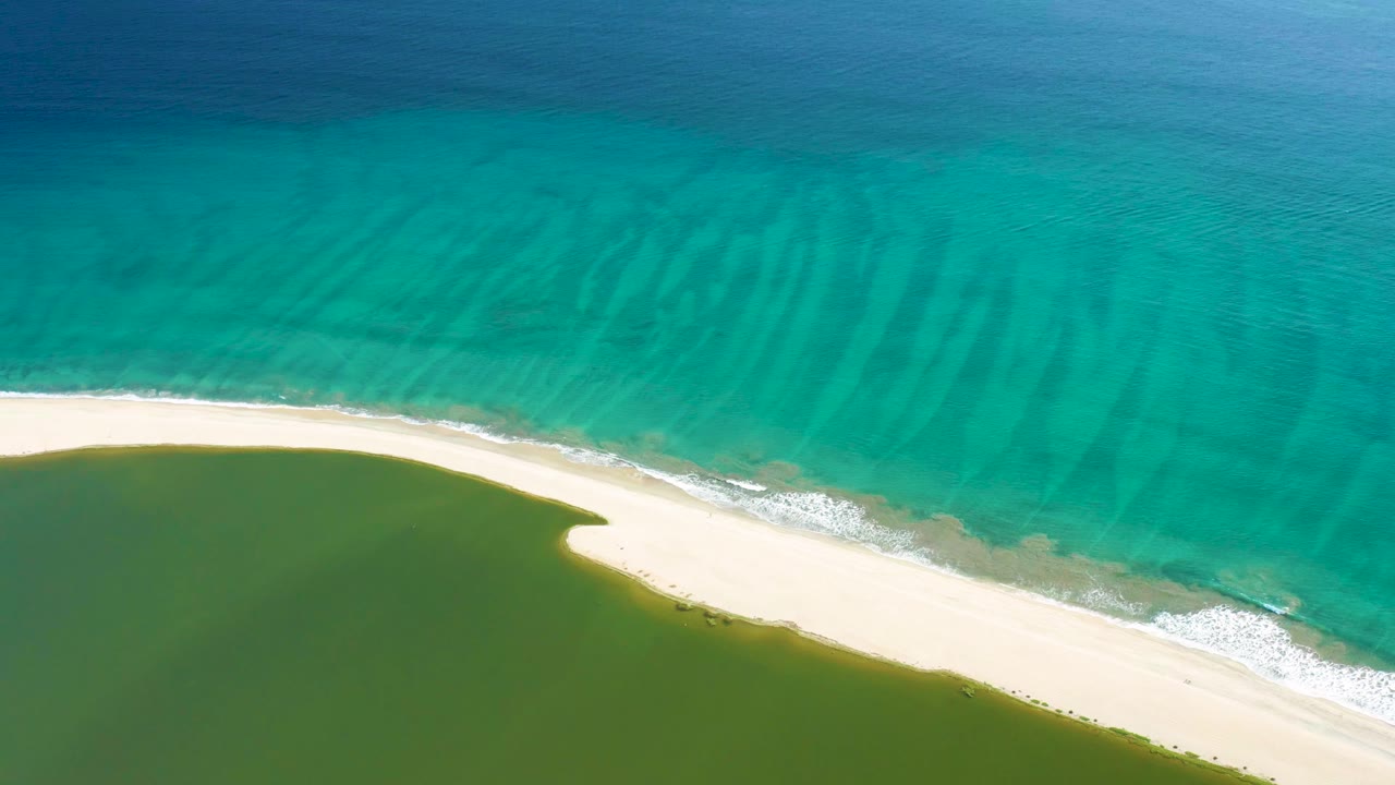 Opening aerial frame of San Jose del Cabo shoreline with turquoise ocean above a pale sandbar and green lagoon below, no visible people.