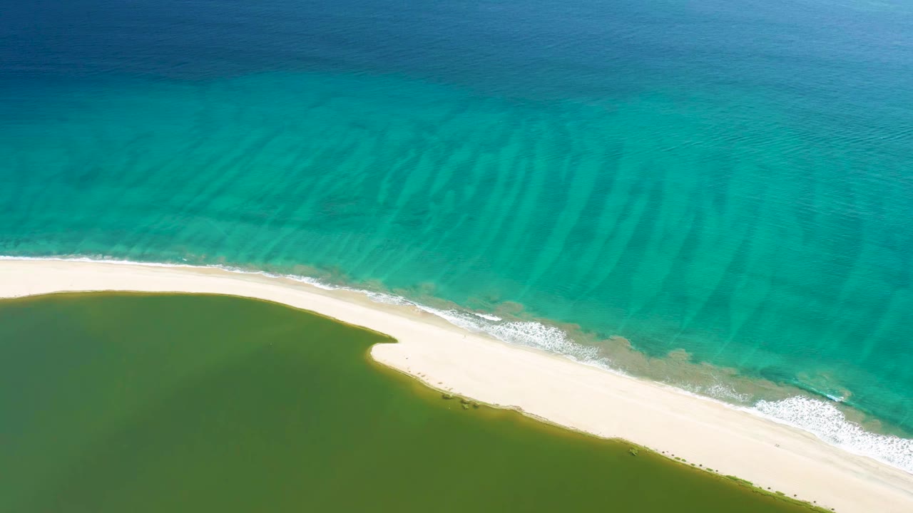 Aerial view one second in shows the San Jose del Cabo sandbar cutting diagonally between turquoise water and the darker green lagoon, no visible people.