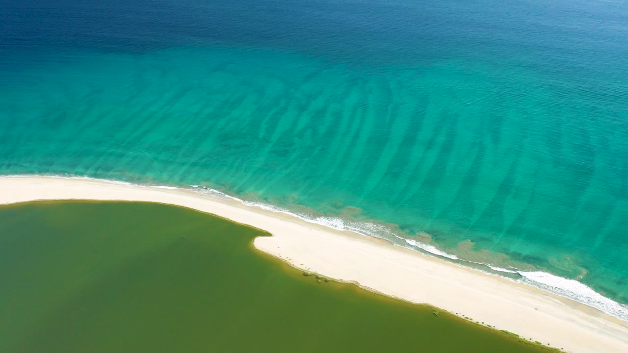 Aerial frame from San Jose Del Cabo showing turquoise ocean above a curved sandy beach and green lagoon below, with no visible people.