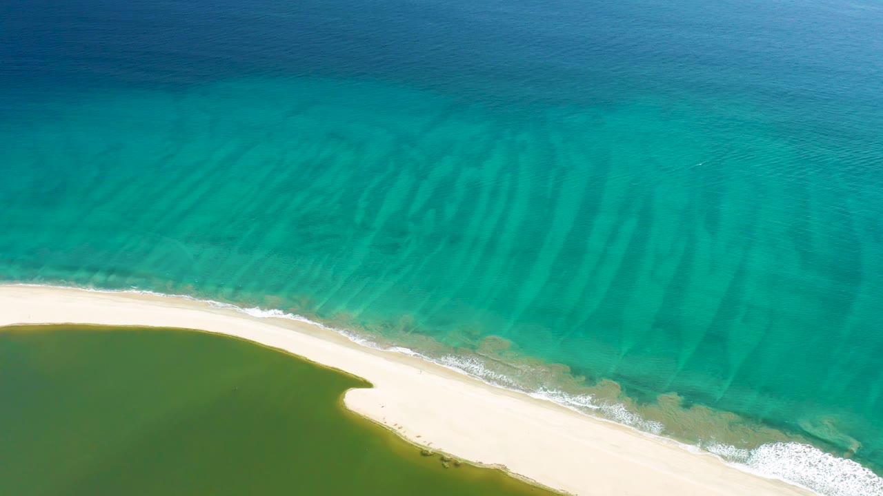 San Jose del Cabo aerial frame with clear turquoise water, visible seabed patterns, a narrow sand strip, and green lagoon water at the lower left, with no visible people.