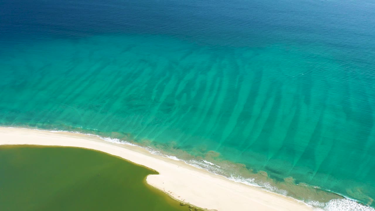 San Jose del Cabo aerial still with turquoise ocean dominating the frame, pale beach along the bottom, green lagoon water at lower left, and no visible people.