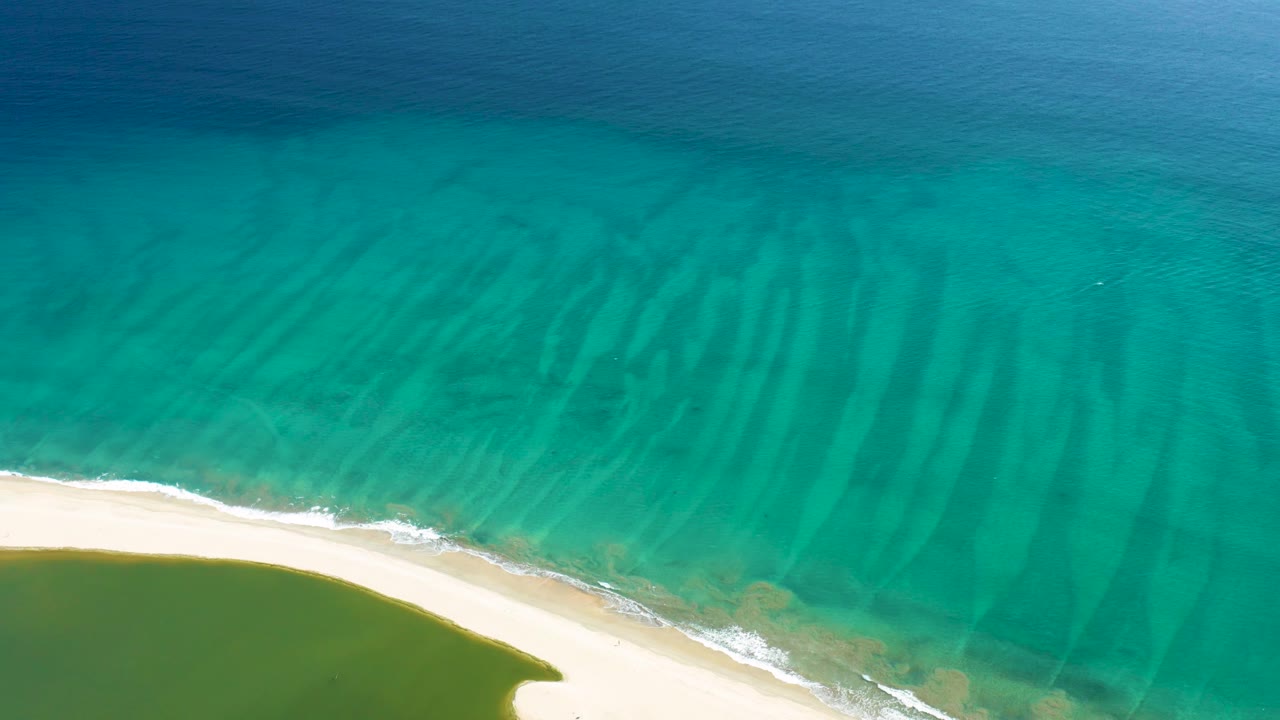 Top-down San Jose del Cabo beach frame with turquoise water filling most of the view, pale sand along the lower edge, a small foam line, and no visible people.