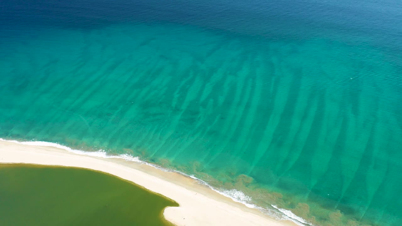 Aerial frame in San Jose del Cabo showing a wider sweep of clear turquoise water above a curving pale beach, green shallows, light foam, and no visible people.