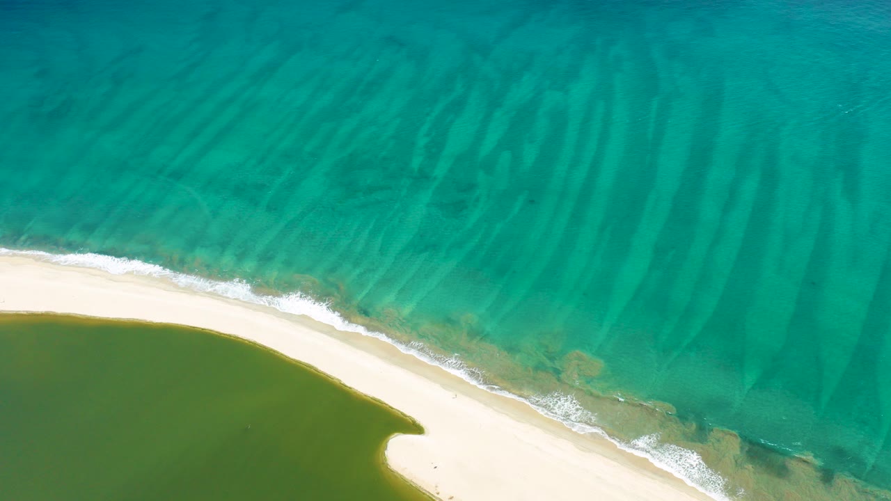 Overhead view in San Jose del Cabo showing turquoise ocean above a curved pale beach and green lagoon water below, with no visible people.