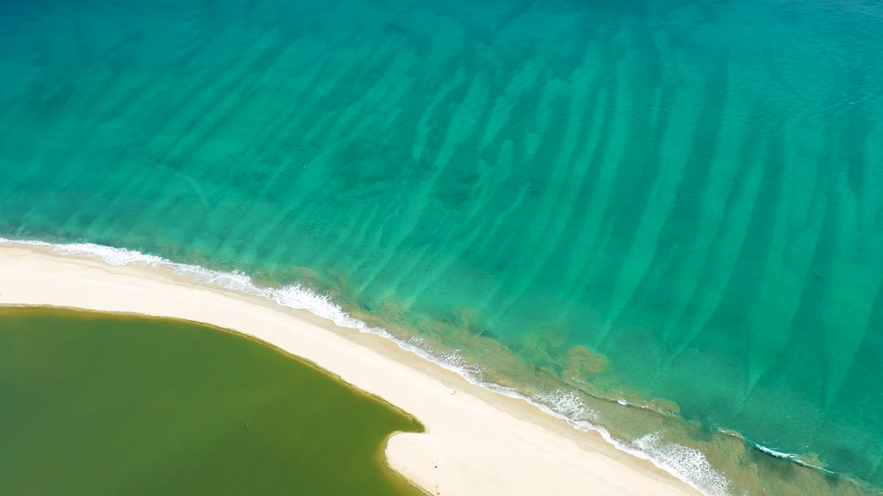 Overhead coastal frame from San Jose del Cabo with blue-green water, diagonal white surf, pale sand, and a small darker wave mark near the right edge, with no visible people.