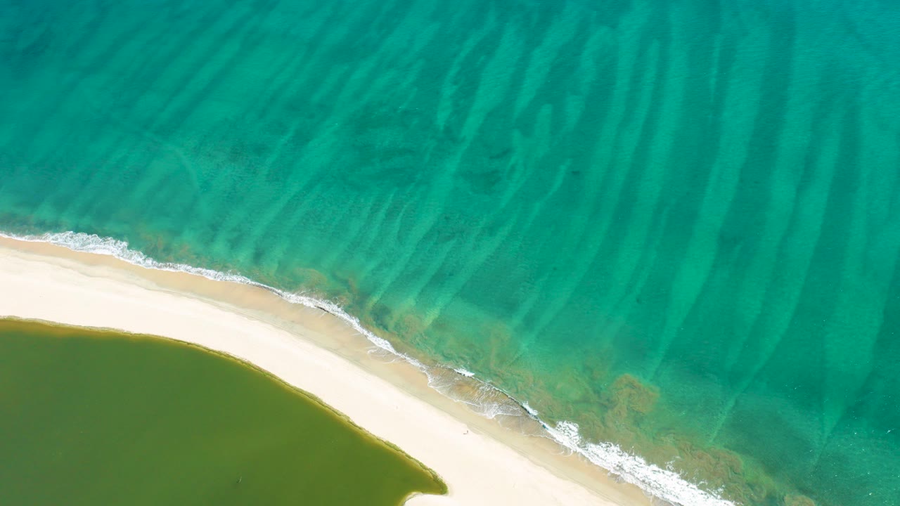 Aerial view in San Jose Del Cabo showing turquoise ocean ripples above a curved pale sand shoreline and green lagoon water below, with no visible people.