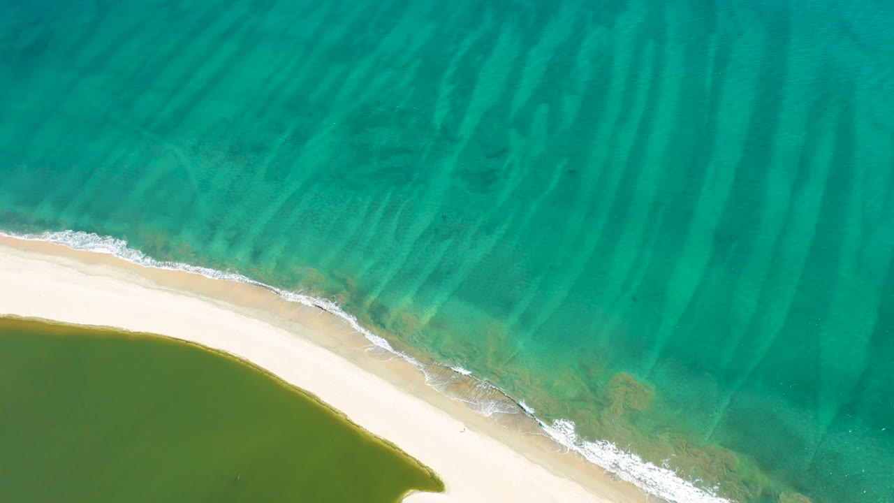 Aerial frame in San Jose Del Cabo with white foam tracing the beach edge as turquoise water covers most of the view and green lagoon water sits at the lower left, with no visible people.