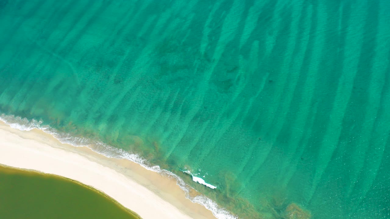 Overhead view in San Jose del Cabo, Mexico, with turquoise rippled shallows, a curved white sand strip at the lower left, small shore foam, and no visible people.