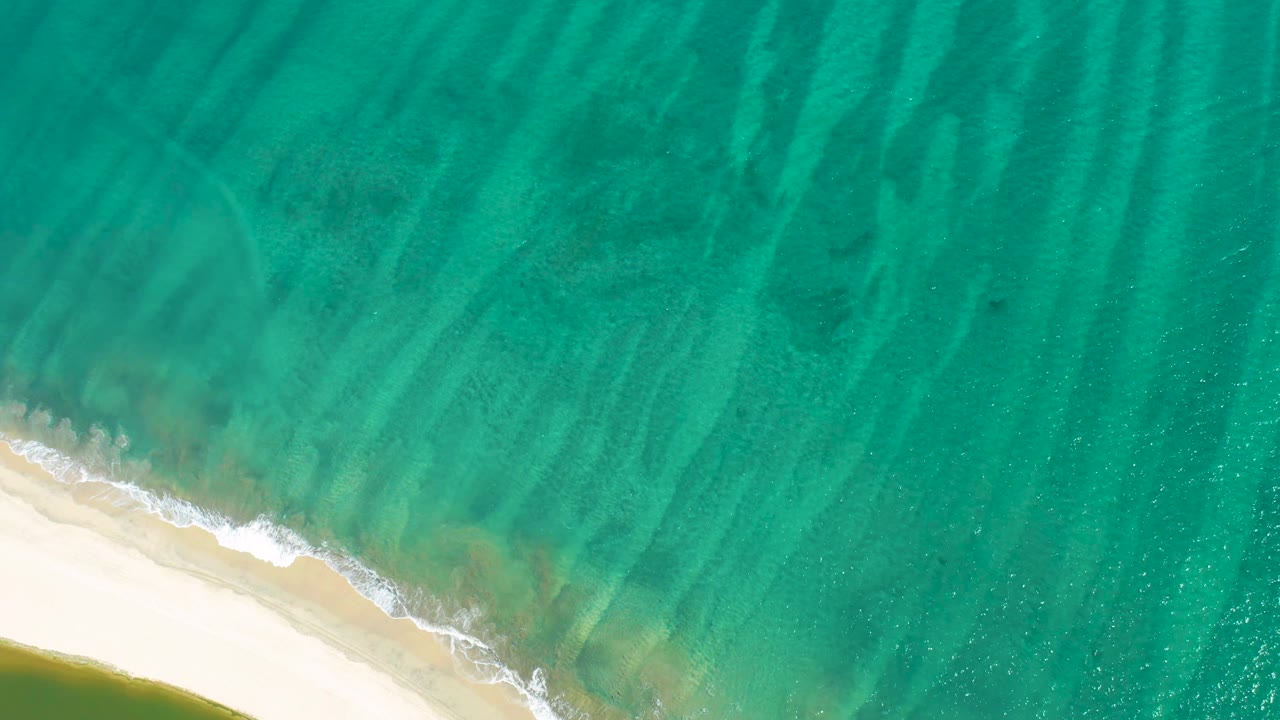 Aerial frame of San Jose Del Cabo surf with pale sand at lower left, translucent green-blue water over seabed streaks, and no visible people.
