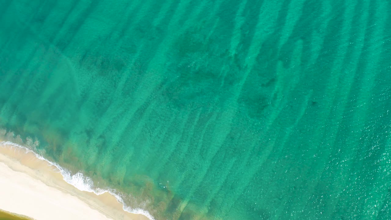 San Jose Del Cabo shoreline seen from above, with white foam tracing the sand at the lower left and clear water revealing soft seabed streaks, no visible people.