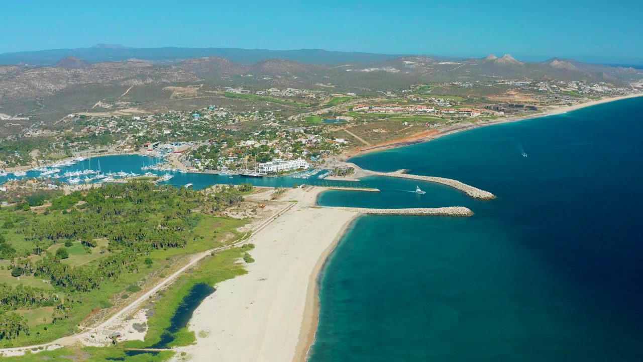 Aerial view over San Jose del Cabo with a pale beach in the foreground, marina basin and breakwater at center, blue sea to the right, dry hills in the distance, and no visible people.