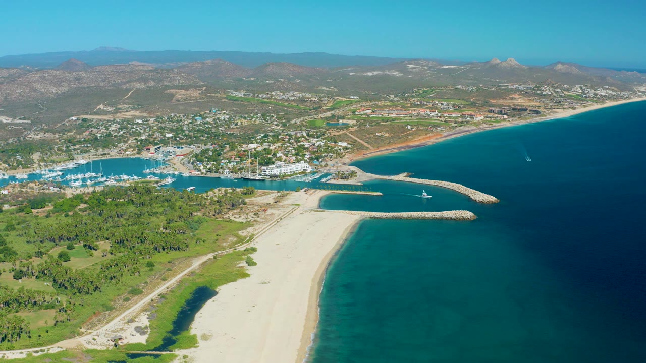 Aerial frame of San Jose del Cabo coastline with the sandy beach filling the lower center, marina entrance and breakwater across the middle, blue water at right, hills across the horizon, and no visible people.