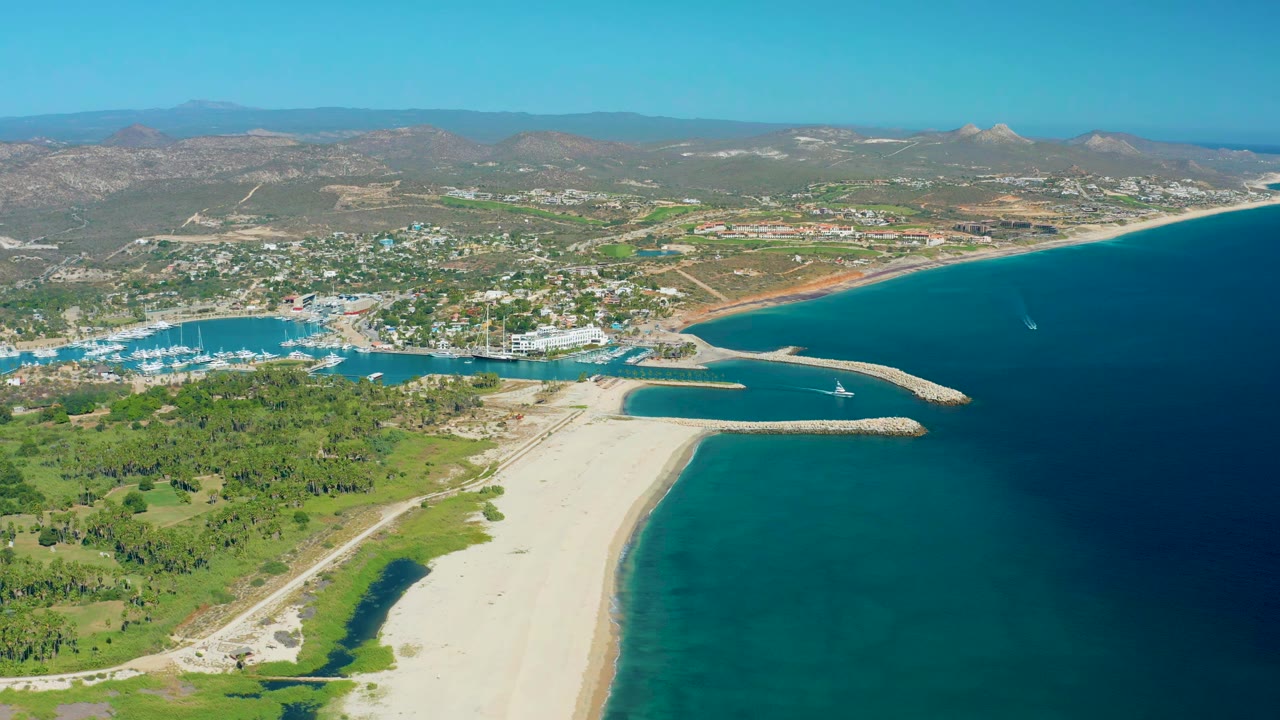 San Jose Del Cabo aerial frame with the marina entrance left of center, a long sandy beach below, darker blue offshore water to the right, mountains in back, and no visible people.