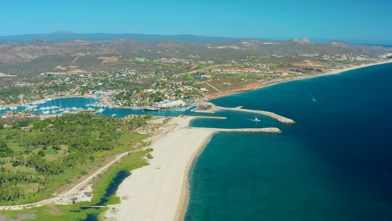 Wide San Jose Del Cabo coastline frame showing the marina and breakwaters across the center, sandy beach below, open blue water on the right, low town development, mountains, and no visible people.