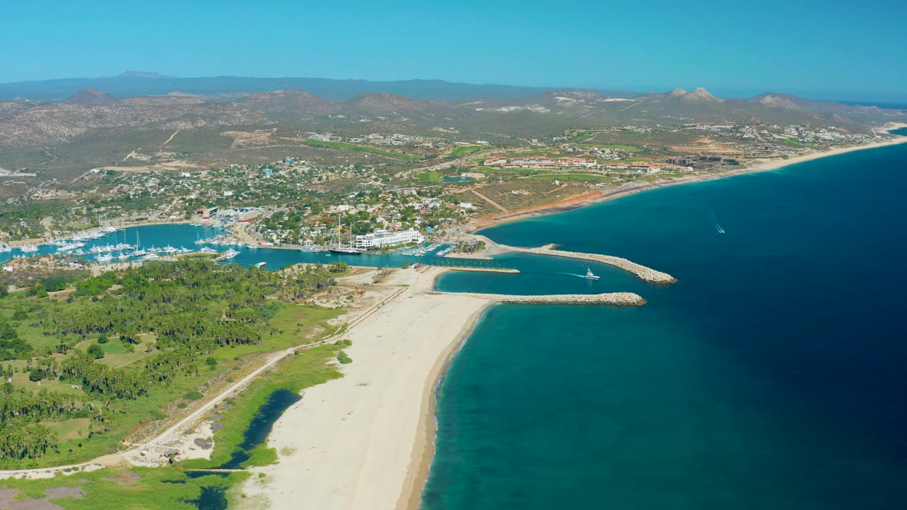 Aerial San Jose Del Cabo view with green foreground land, pale beach, marina docks at left, breakwaters near center, deep blue water at right, distant mountains, and no visible people.