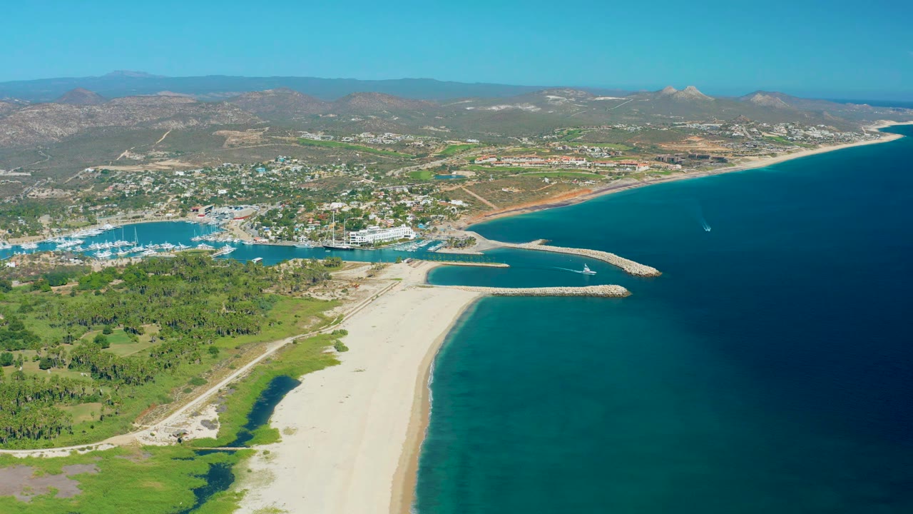 Aerial view of San Jose del Cabo with the beach running up the frame, the marina basin on the left, deep blue water on the right, and no visible people.